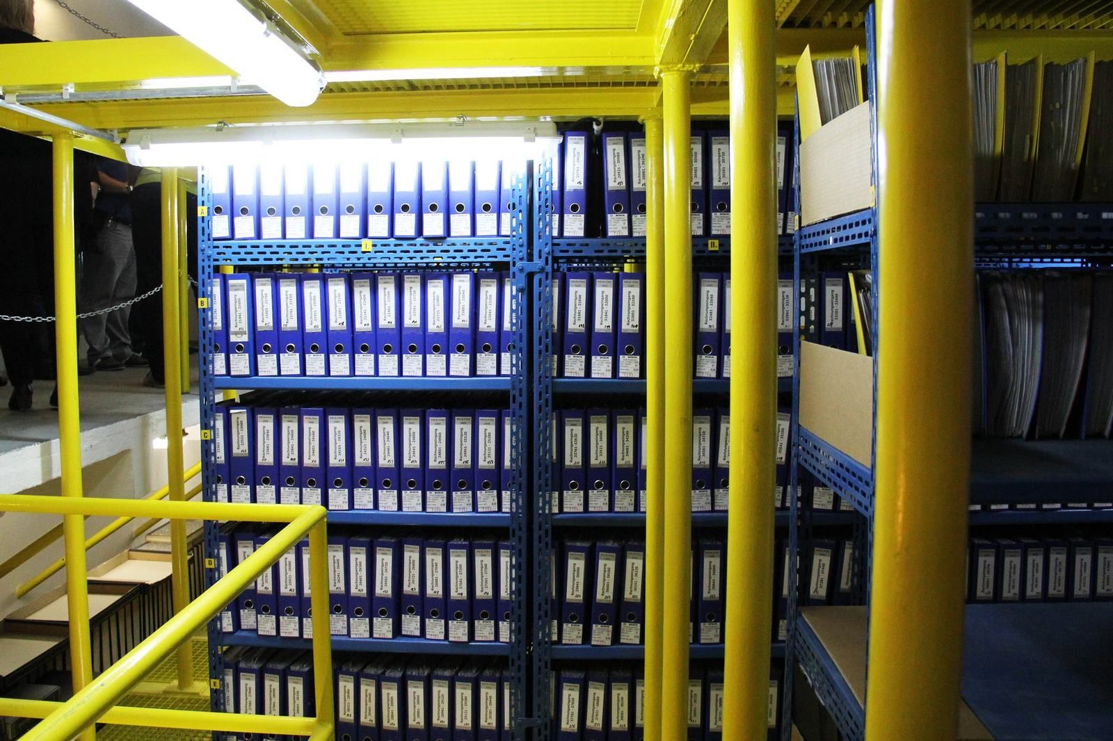 Žlutá ocelová patra a modré regály se šanony ve spisovně ZÁSOBY